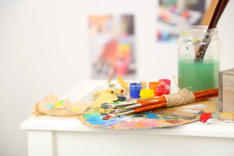Una mesa con una paleta de pintor y varios pinceles manchados de pintura de colores, en una clase de dibujo y pintura.